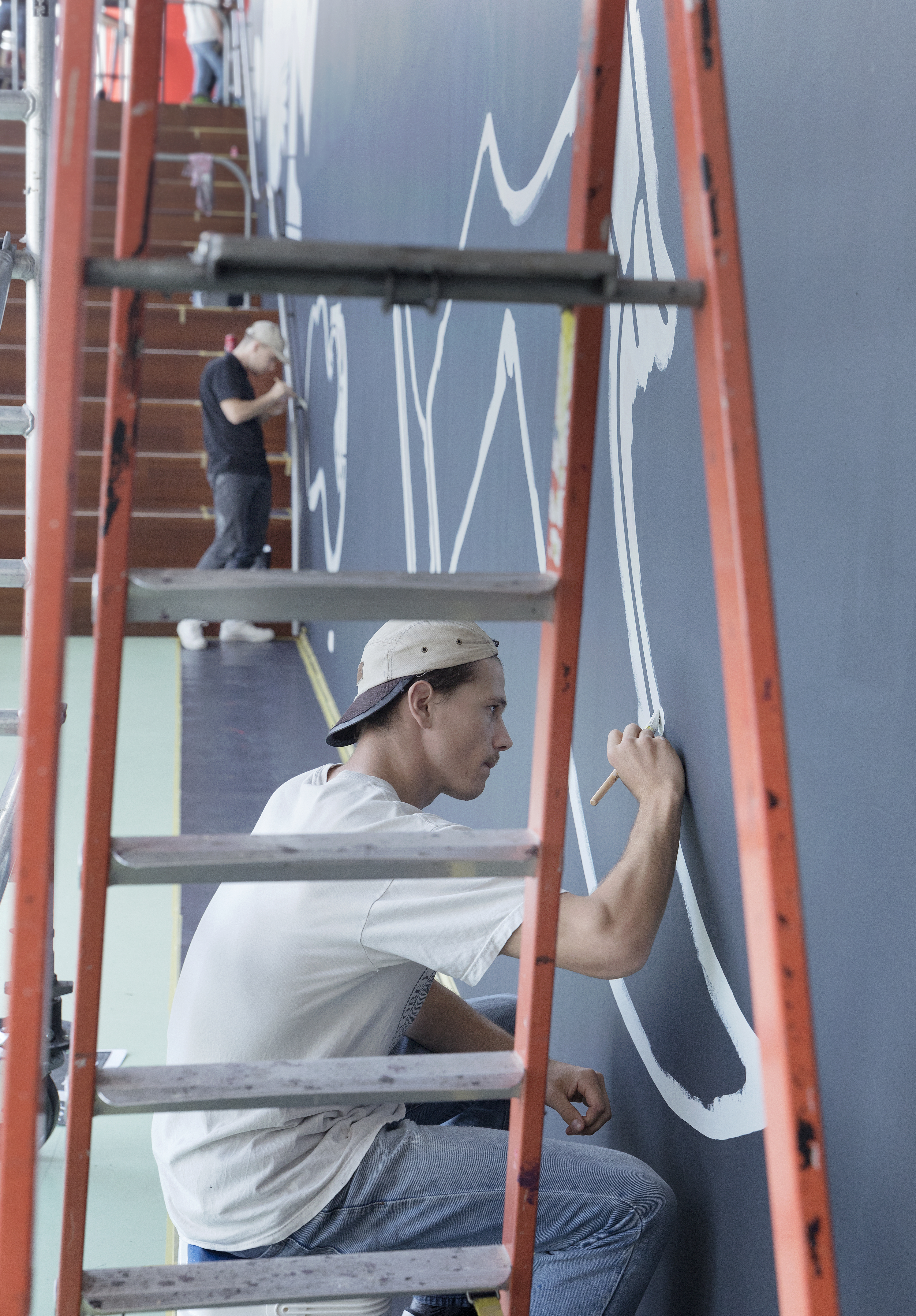 een assistent en Lennard Kok werken aan de muurschildering onder een steiger